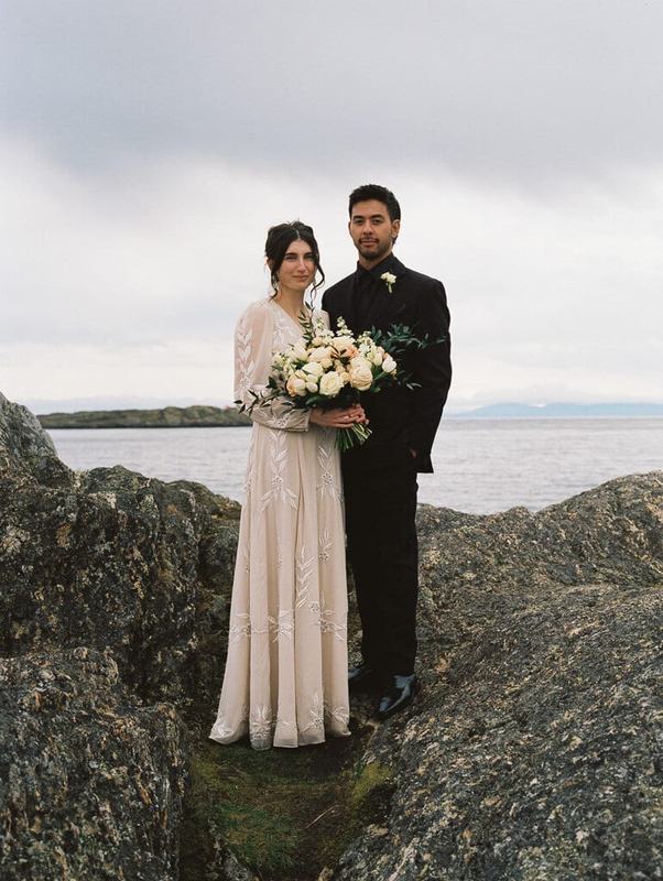 Bride and groom with white roses standing on coastal rocks under grey skies