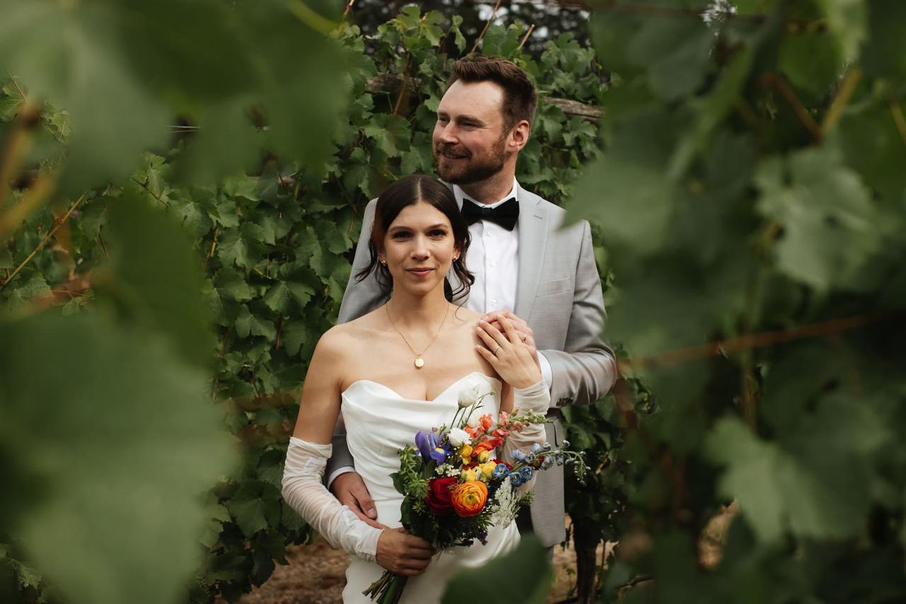 Bride and groom holding colorful bouquet stand before vineyard foliage at Church and State Winery
