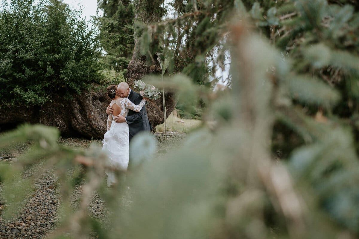 Couple embracing by mossy boulders, framed through evergreen branches