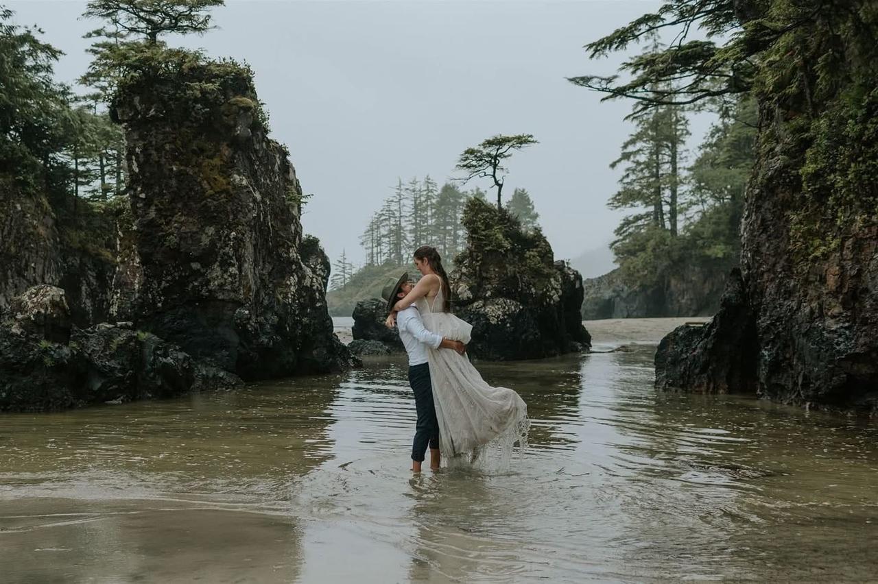 Couple embracing in shallow water during rain with windswept tree and forest on Vancouver Island