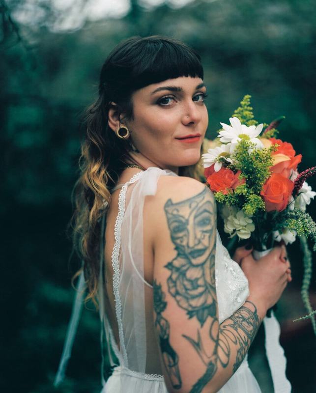 Bride with blunt bangs and tattooed arm holding an orange bouquet