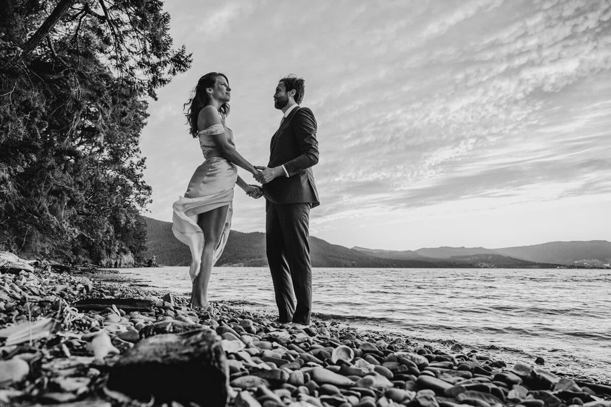 Couple holding hands on a pebble beach, dress catching the wind
