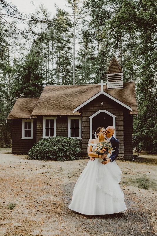 Groom nuzzling his bride outside a shingled woodland chapel