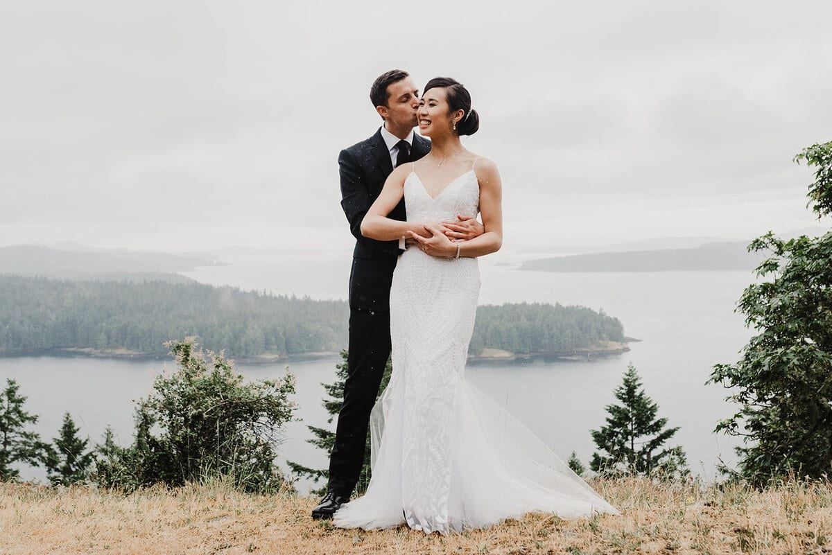 Groom kissing the bride's cheek on a bluff above island waters
