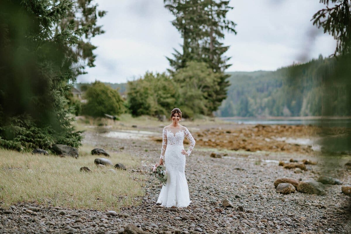 Bride in long-sleeved lace crossing a stony inlet shoreline