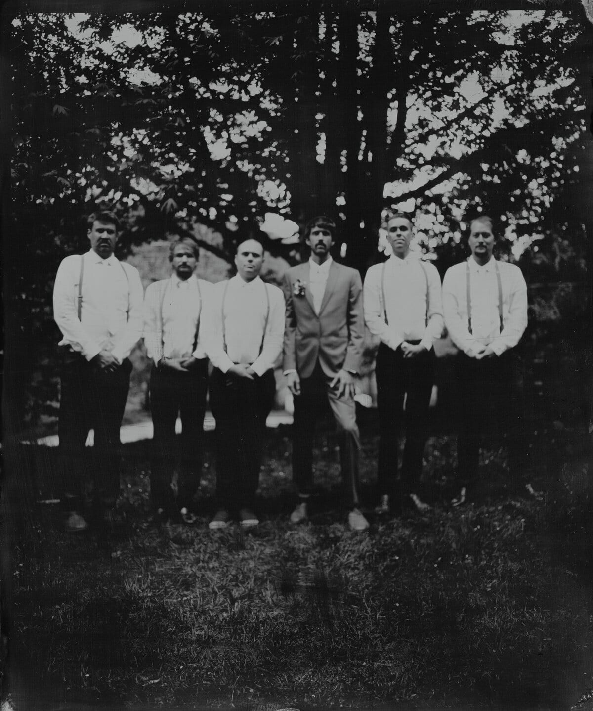 Six men in suspenders and shirtsleeves posing under shade trees
