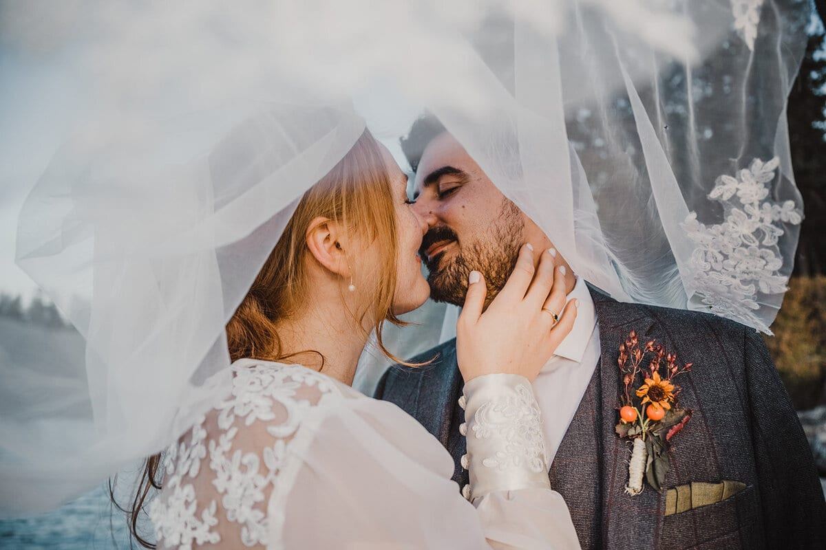 Couple kissing beneath a sheer veil, orange boutonniere visible