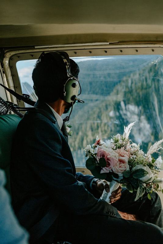 Groom in a helicopter headset carrying the bouquet over mountain cliffs