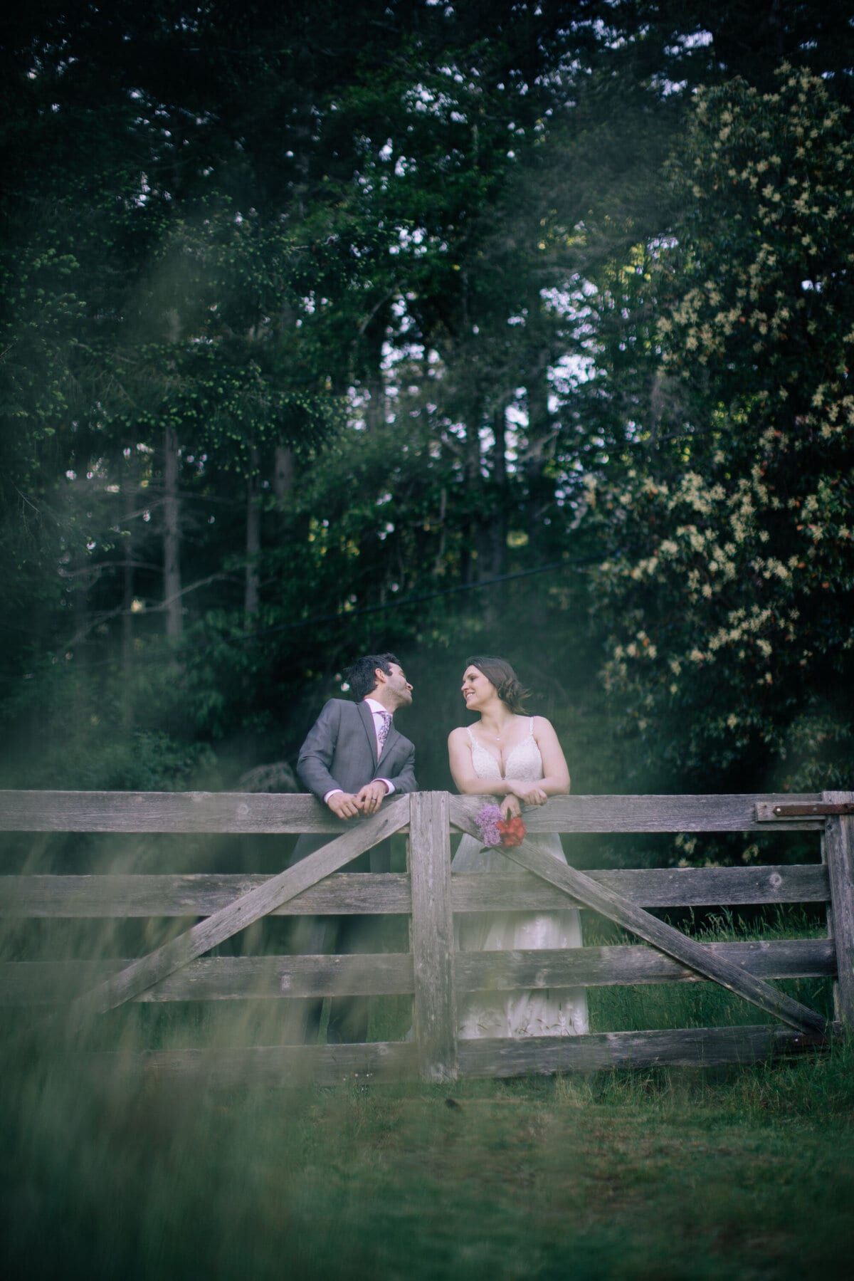 Newlyweds trading glances across the top rail of a gate