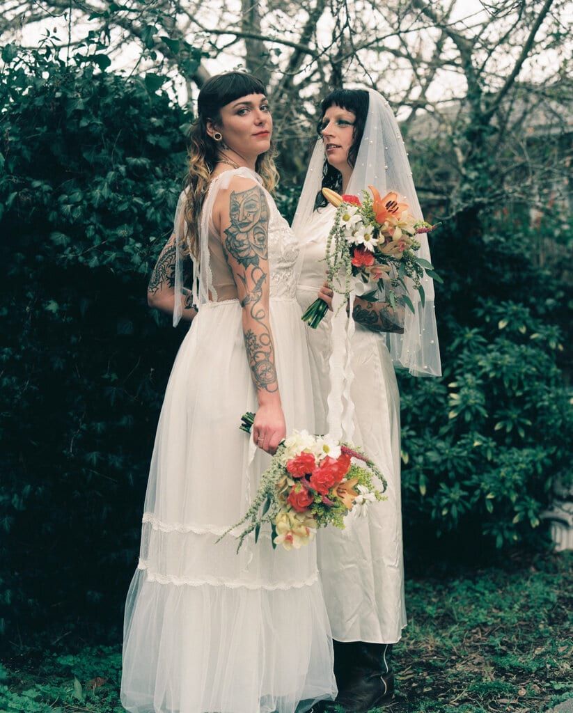 Two tattooed brides holding bright bouquets in an evergreen garden