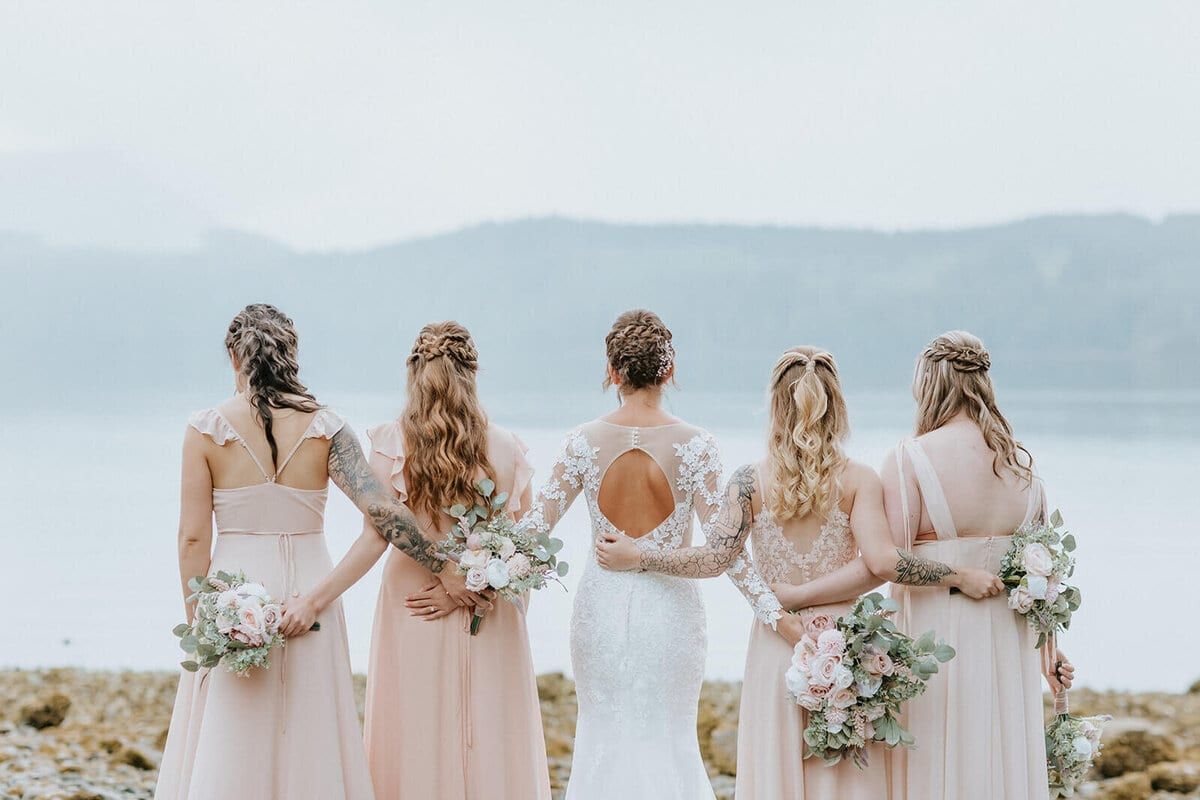 Bride and bridesmaids in blush linking arms before a misty lake