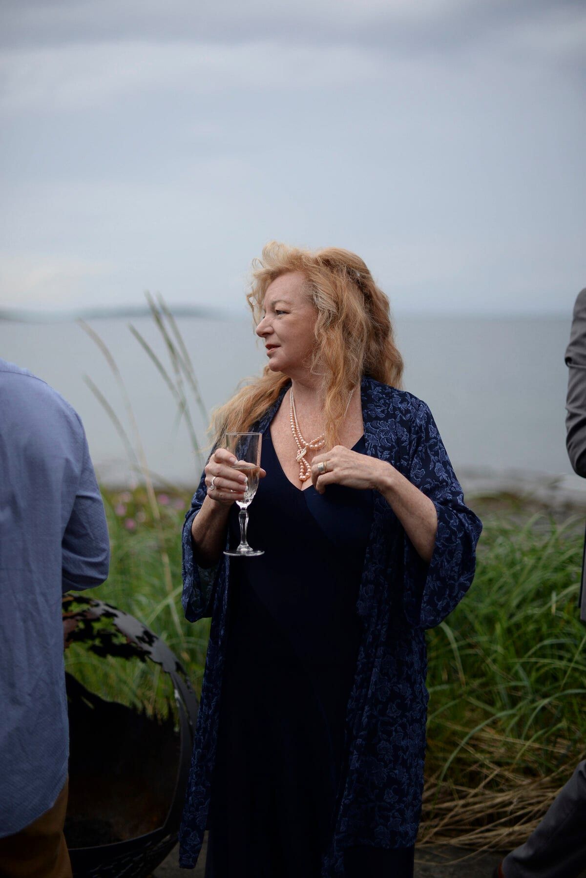 Red-haired guest in a navy shawl holding champagne by the shore