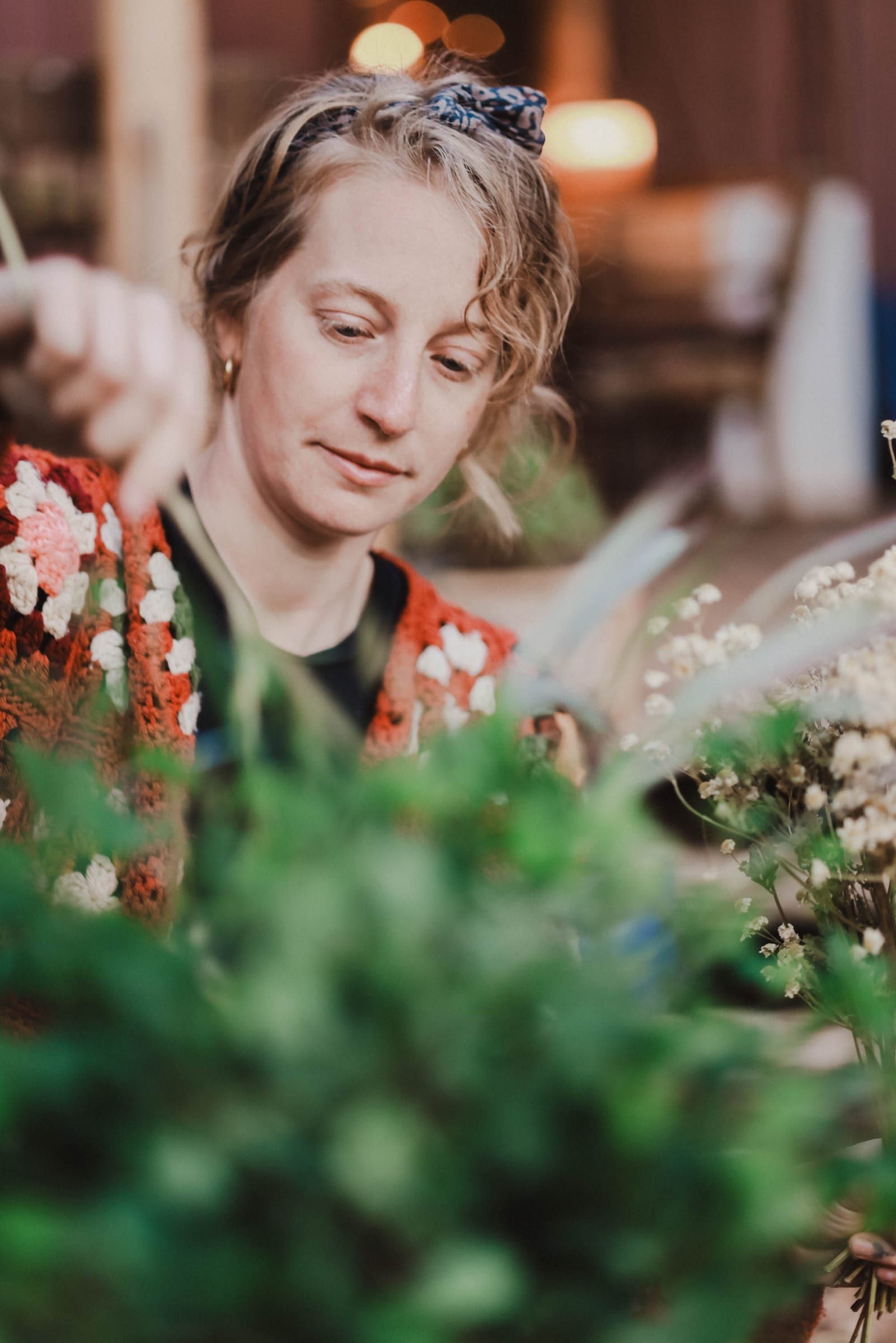 Woman with wavy blonde hair and blue patterned headband gazes down at red and white wildflowers, soft bokeh lights glowing behind her