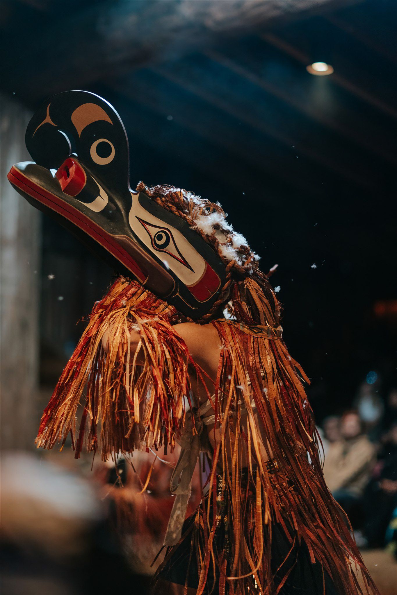 Indigenous carved paddle with eye design rests against dark water, adorned with red and orange cedar fringe in golden sunlight