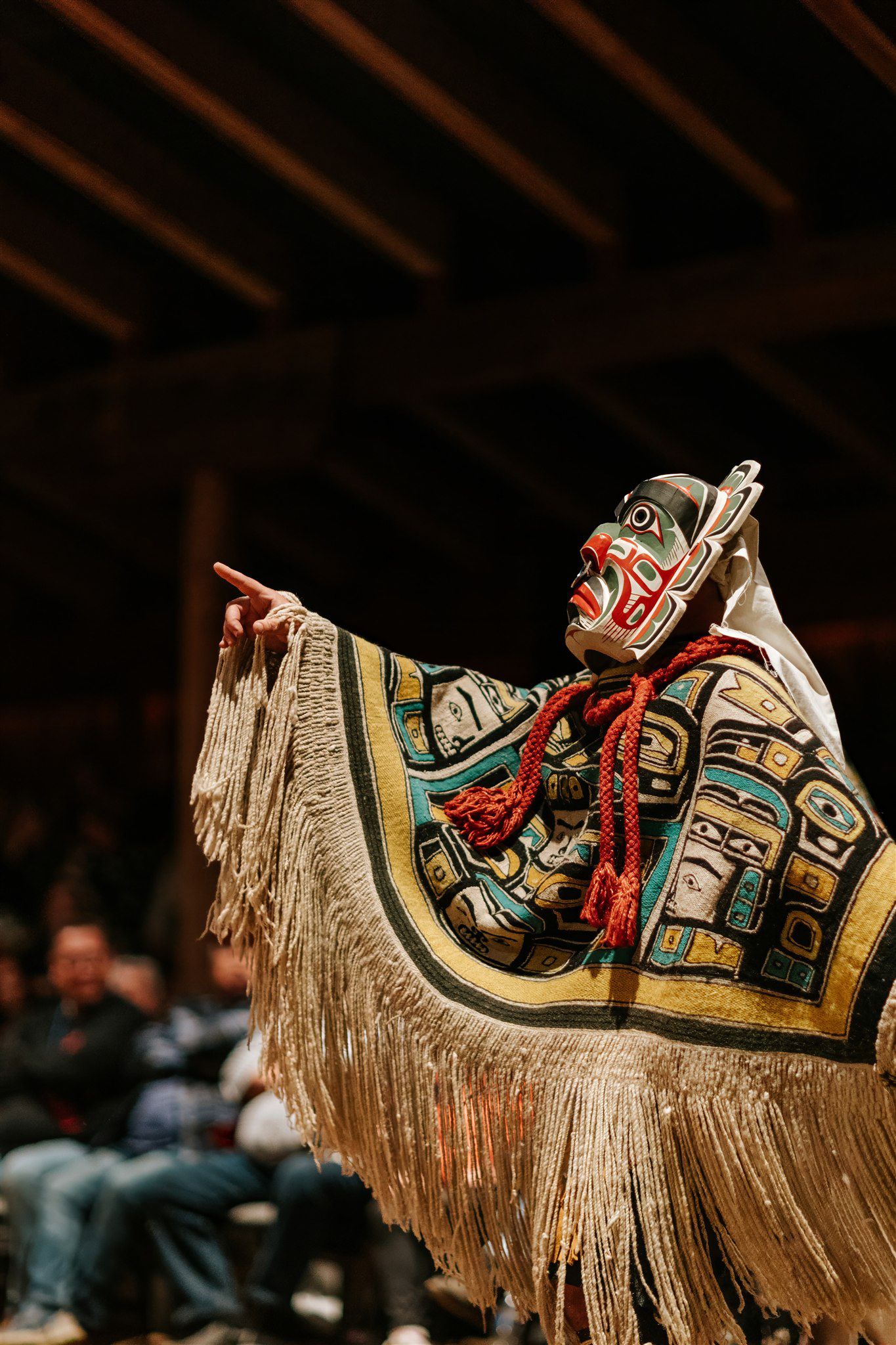 Ornate button blanket with cream fringe and red braided ties draped against dark background, detail of Indigenous regalia
