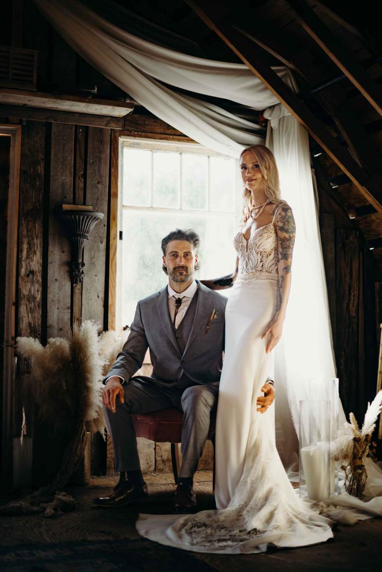 Groom in grey suit seated beside bride in white dress and veil, sunlit windows behind, wooden beams overhead
