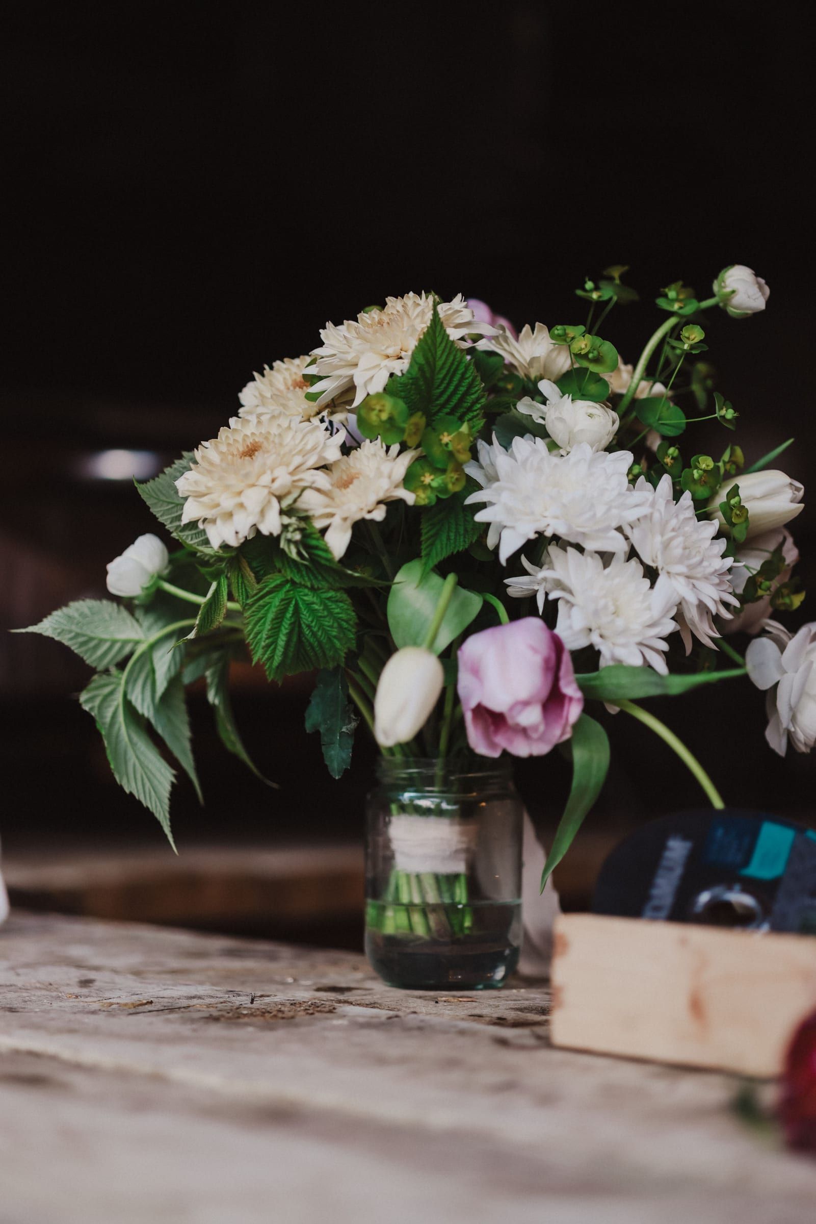 Cream and blush dahlias with green hypericum berries and textured foliage arranged in a clear glass vase against a dark background