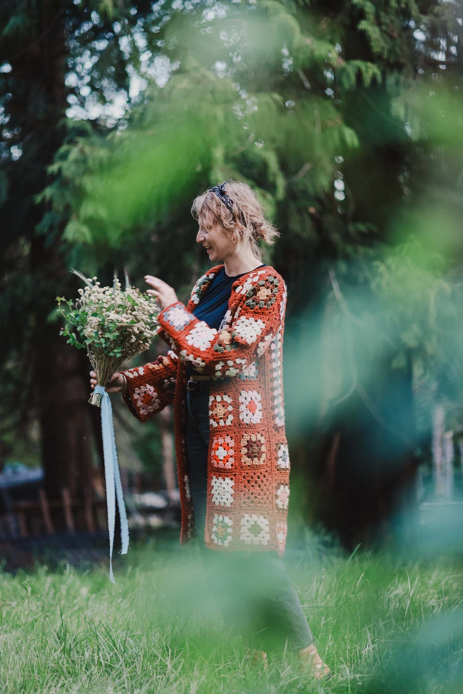 Person in ornate crocheted cardigan with orange and cream granny squares stands smiling among lush green foliage and white flowering shrubs