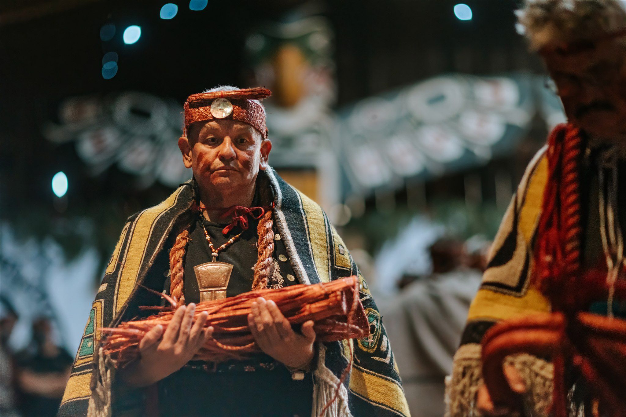 Elder in traditional regalia holds red-wrapped gift, warm indoor lighting with blurred figures behind